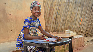 Ein Mädchen mit traditioneller, bunter Kleidung sitzt an einem kleinen Tisch. Vor ihr liegt ein Heft und sie hält einen Stift in der Hand. Sie lächelt in die Kamera.